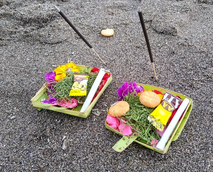 Two Balinese canang offerings with flowers and incense on dark sand