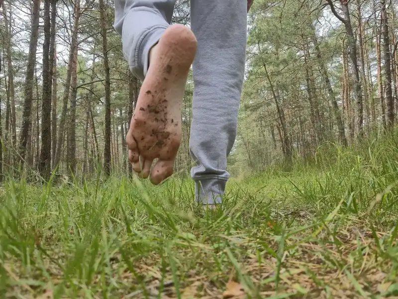 A barefoot person walks away through grass in a pine wood, one soil-stained sole lifted toward the camera.
