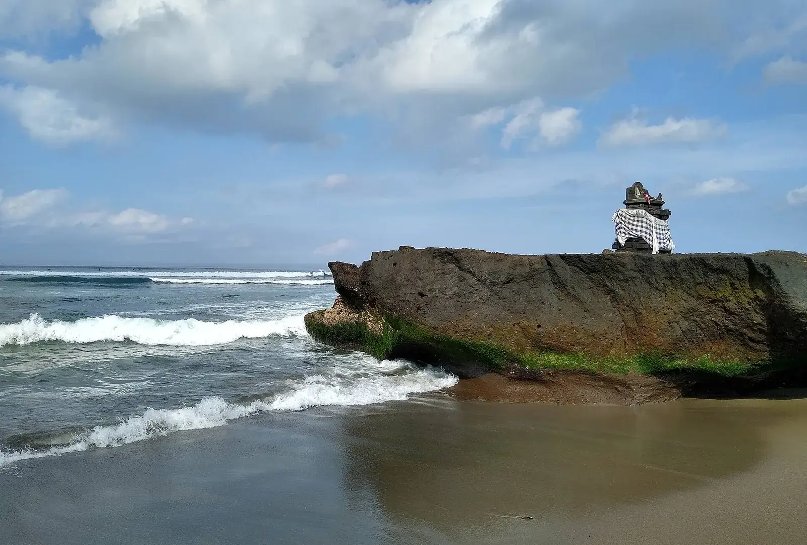 A small stone shrine wrapped in black-and-white checked cloth stands on a weathered rock above the sand as waves break along the shore in Bali.