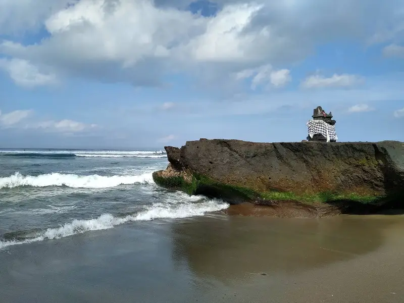 A small stone shrine wrapped in black-and-white checked cloth stands on a weathered rock above the sand as waves break along the shore in Bali.