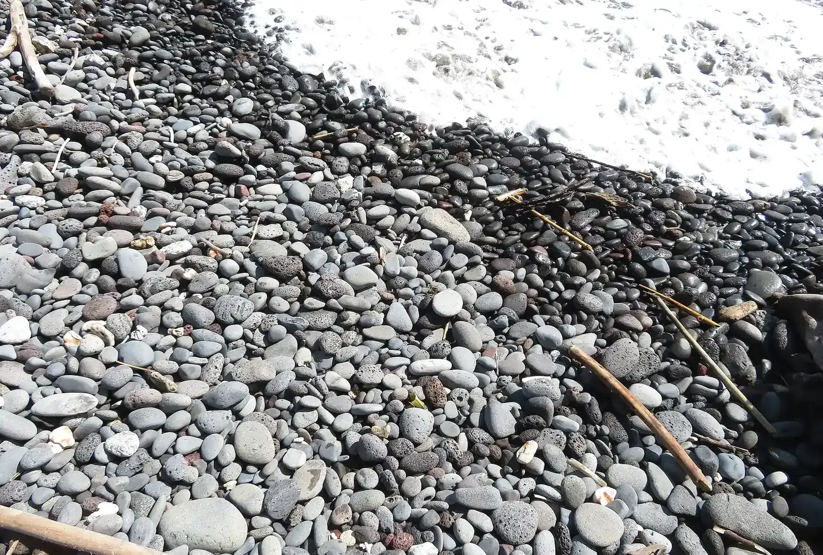 Rounded grey and black pebbles on a shoreline with white sea foam washing over them.