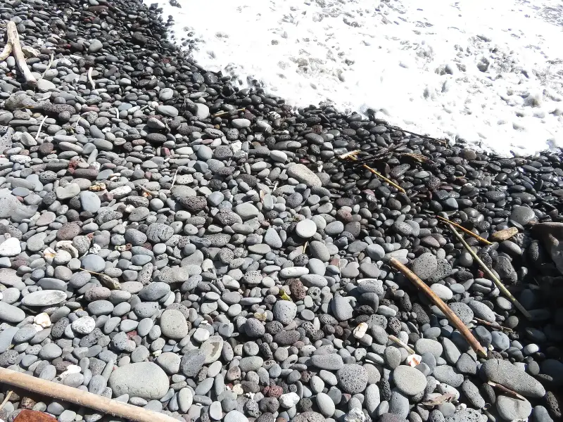 Rounded grey and black pebbles on a shoreline with white sea foam washing over them.